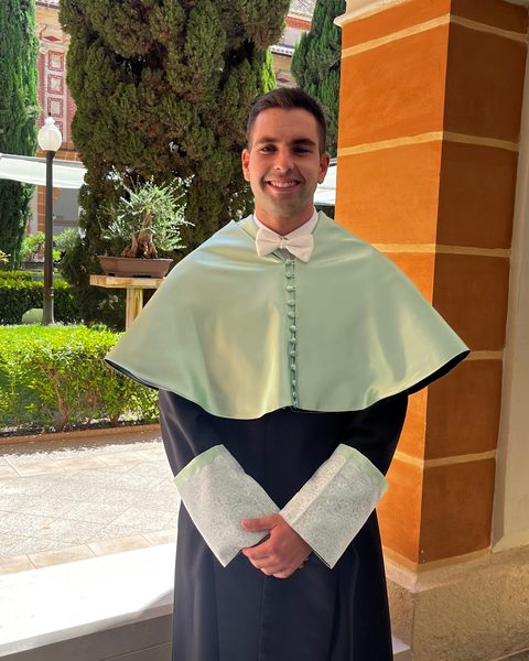 Antonio Vélez Alcázar sonríe con la vestimenta académica de doctor, en un claustro con jardín.